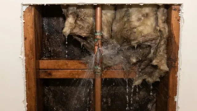 Opened wall with water spraying from a burst pipe through wet insulation