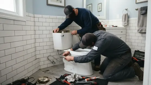 Two plumbers working on a toilet tank in a tiled bathroom